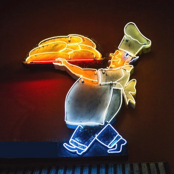 Vintage neon sign of a chef carrying a tray of bread, illuminated in white, yellow, and red colors. The sign is mounted on a dark wall, adding a nostalgic and charming ambiance to the space, ideal for a bakery or restaurant.