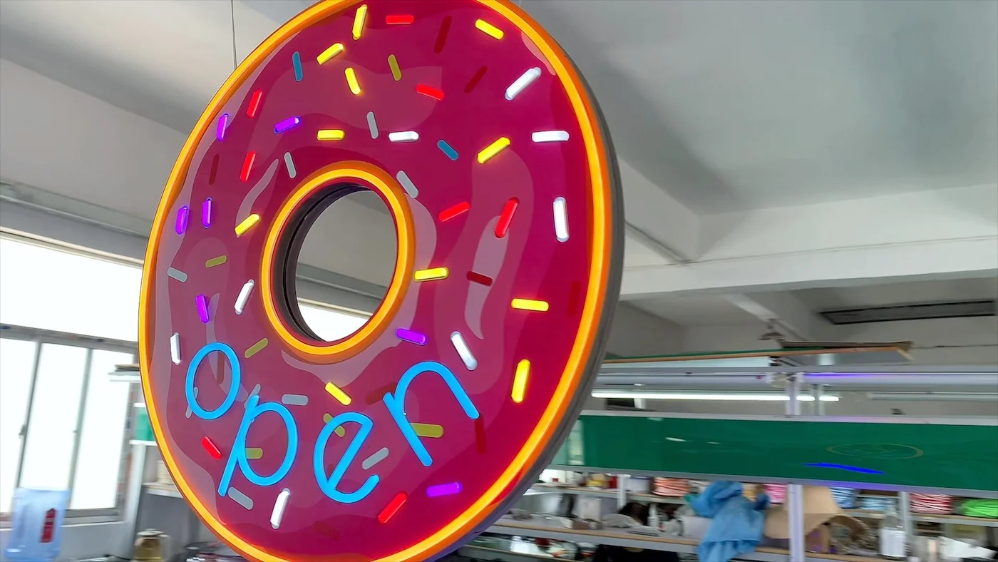 Neon sign in the shape of a pink frosted donut with colorful sprinkles, displaying the word "open" in blue letters. The sign is illuminated and hanging in a workshop environment, showcasing a vibrant and playful design ideal for a bakery or donut shop.