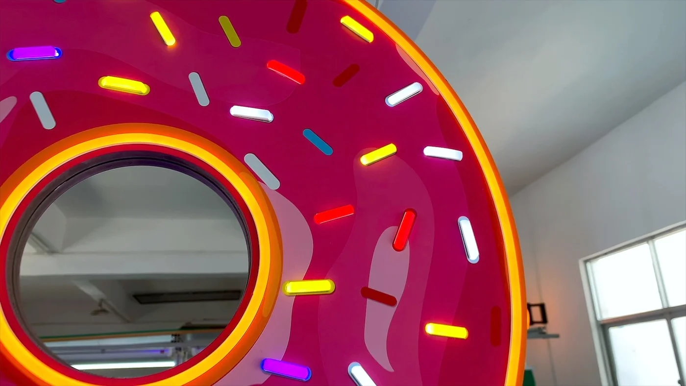 Neon sign in the shape of a pink frosted donut with colorful sprinkles, displaying the word "open" in blue letters. The sign is illuminated and hanging in a workshop environment, showcasing a vibrant and playful design ideal for a bakery or donut shop.