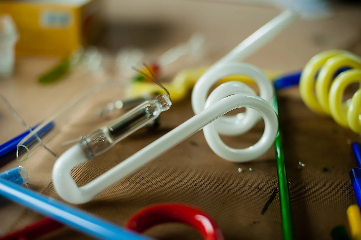 Various glass tubes and components for neon signs, including a white spiral tube and colorful straight tubes, spread out on a workbench. The image highlights the materials used in the neon sign-making process.