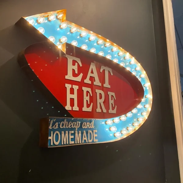 A vintage neon sign on a wall with a large blue arrow outlined with lightbulbs. The arrow points to the words "EAT HERE" in bold white letters on a red background. Below, a smaller blue section reads "It's cheap and HOMEMADE" in white text.