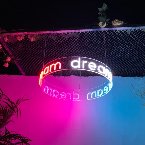 Circular neon sign with the word "dream" repeated, half in red and half in blue, hanging from a ceiling with a wire grid decorated with flowers. The sign casts a colorful glow against the pink and blue illuminated walls, creating a dreamy and artistic atmosphere.