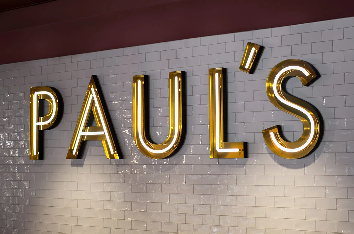 Illuminated neon sign reading "PAUL'S" mounted on a tiled wall inside a restaurant. The sign features large, bold letters with a yellow and white glow, enhancing the upscale ambiance of the venue. The sign is situated above a hallway leading to other areas of the restaurant.