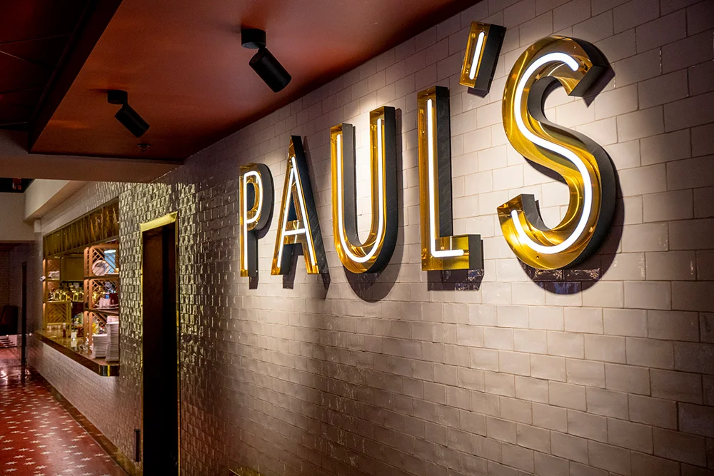 Illuminated neon sign reading "PAUL'S" mounted on a tiled wall inside a restaurant. The sign features large, bold letters with a yellow and white glow, enhancing the upscale ambiance of the venue. The sign is situated above a hallway leading to other areas of the restaurant.