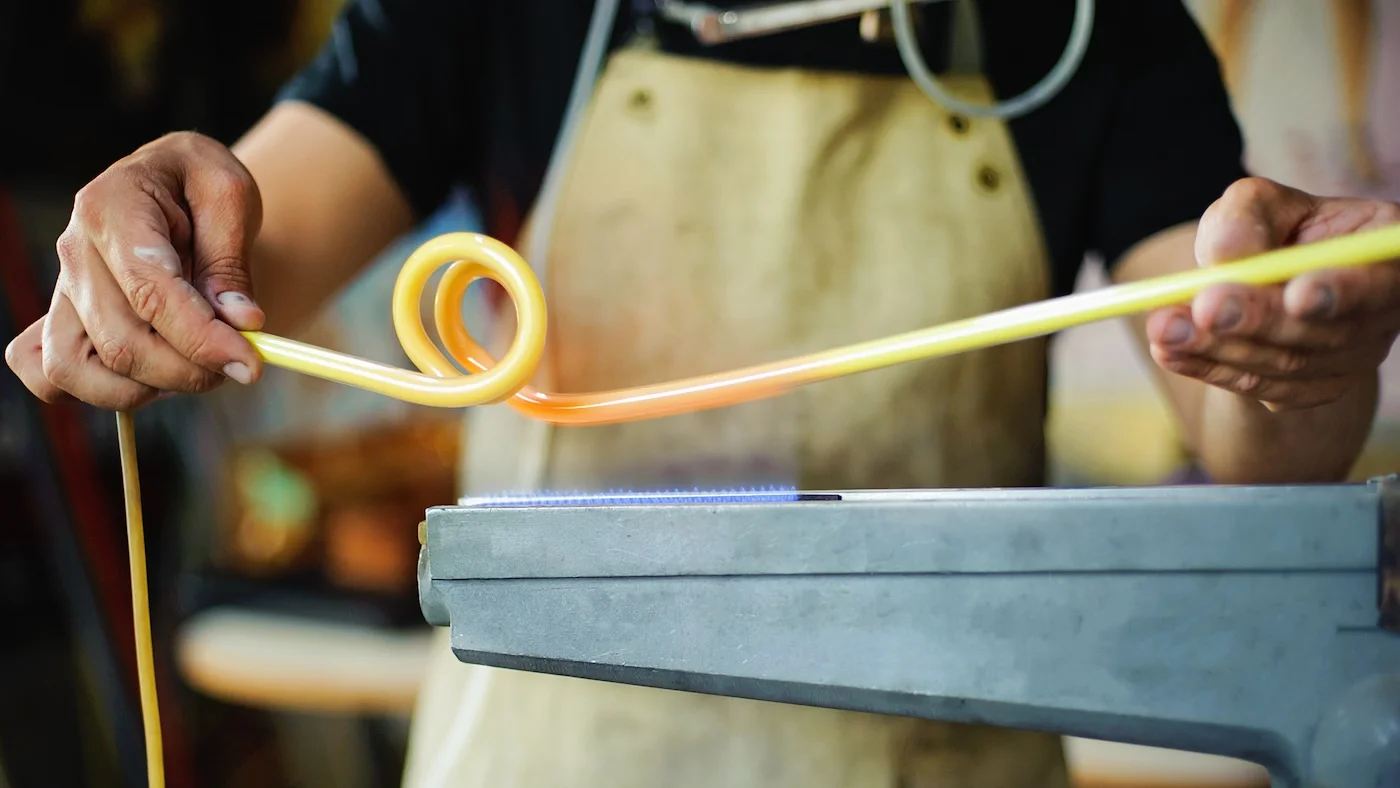 Close-up of a craftsman bending a heated glass tube into a spiral shape over a flame, preparing it for a neon sign. The worker wears an apron, emphasizing the precision and skill involved in neon sign creation.