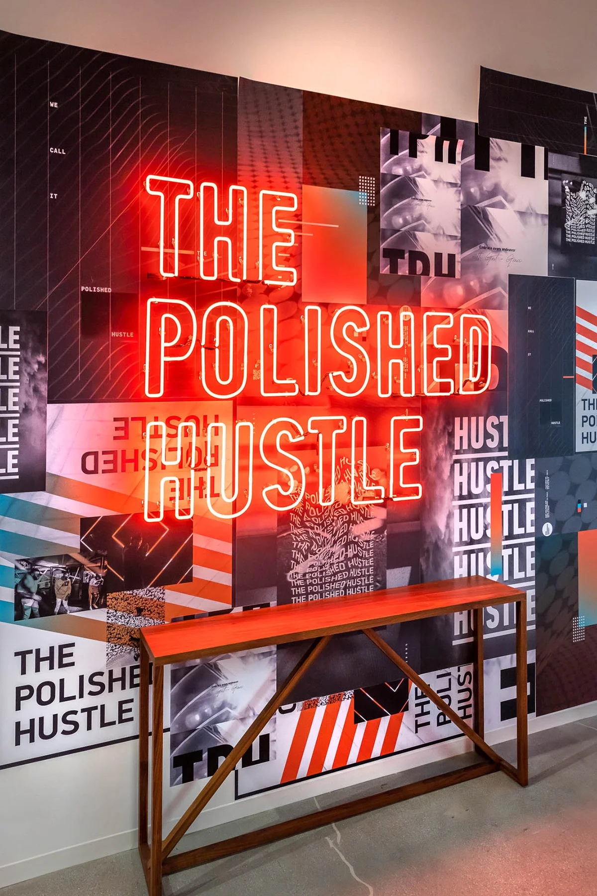 Neon sign reading "THE POLISHED HUSTLE" in red, mounted on a wall covered with various posters and graphics. The sign illuminates a modern and dynamic space, perfect for a creative or business setting.