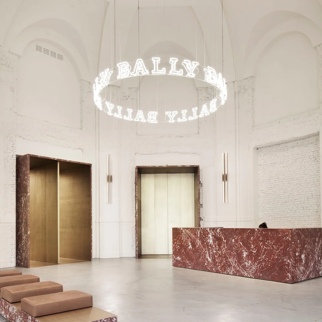 Circular neon sign with the word "BALLY" repeated, in white, hanging from the ceiling of a modern, minimalist lobby.