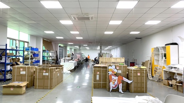 A spacious sign manufacturing facility with large cardboard boxes and packaging materials.   There are workers in the background, various equipment and materials related to signage production.