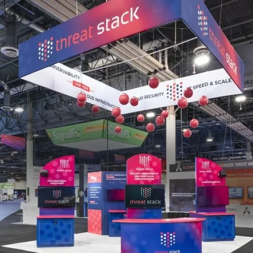 Booth for trade show, featuring large, illuminated signage with the company's logo and branding colors of blue, red, and white. The booth includes multiple display stations and hanging red spheres, creating a visually engaging setup.