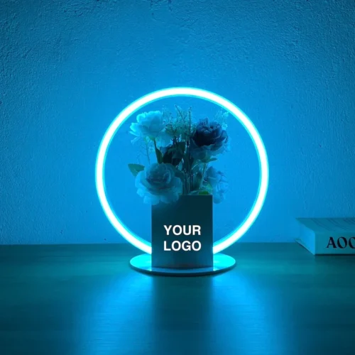 Decorative neon sign featuring a circular blue neon light surrounding a vase with flowers. The vase has a placeholder text "YOUR LOGO," suggesting customizable branding options. The setup is placed on a wooden surface against a plain wall, creating a minimalist and modern look.