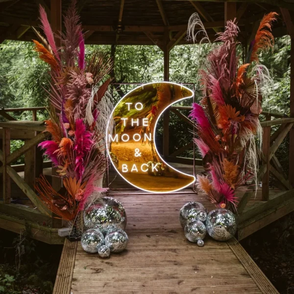 Event signage featuring a crescent moon-shaped neon sign with the words "TO THE MOON & BACK" in white neon. The sign is flanked by vibrant floral arrangements with pink, orange, and beige pampas grass, adding a bohemian touch. Silver disco balls are placed at the base, enhancing the festive atmosphere.