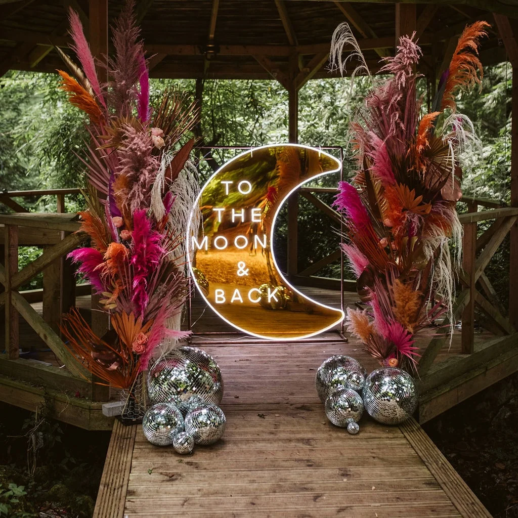 Event signage featuring a crescent moon-shaped neon sign with the words "TO THE MOON & BACK" in white neon. The sign is flanked by vibrant floral arrangements with pink, orange, and beige pampas grass, adding a bohemian touch. Silver disco balls are placed at the base, enhancing the festive atmosphere.