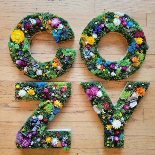 Decorative sign spelling "COZY" made from moss-covered letters adorned with colorful artificial flowers and small butterflies, placed on a wooden floor.