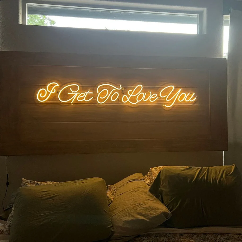 Yellow cursive neon sign "I Get To Love You" on a wooden background above a cozy bed.