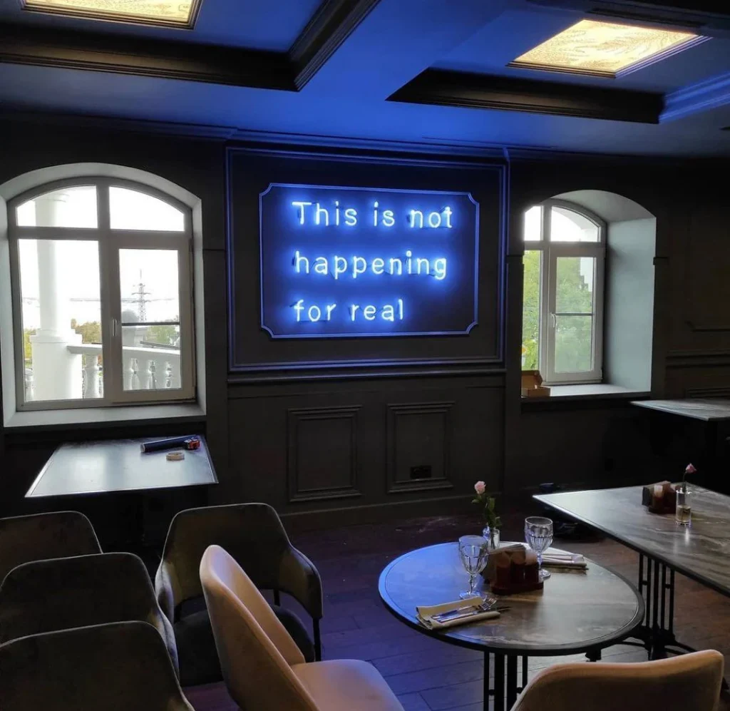 A restaurant interior with a blue neon sign that reads "This is not happening for real" on a dark wall.