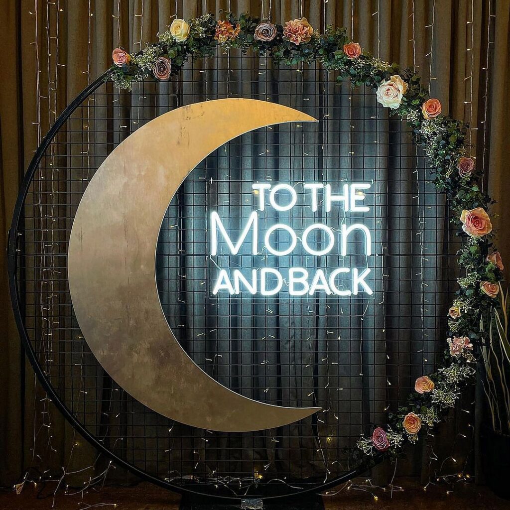 Crescent moon and circular neon sign with "TO THE MOON AND BACK" text, framed by a floral garland and string lights.