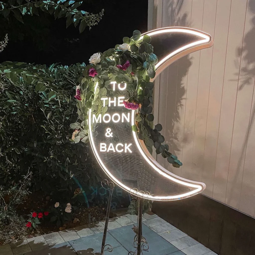 Crescent moon-shaped neon sign with "TO THE MOON & BACK" text, decorated with greenery and pink flowers against an outdoor backdrop.