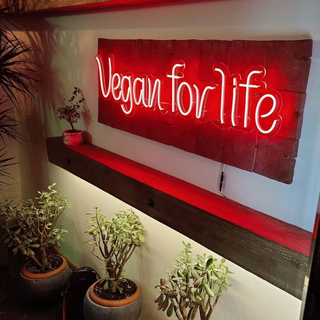 Red cursive neon sign "Vegan for life" on a rustic wooden background with potted plants below.