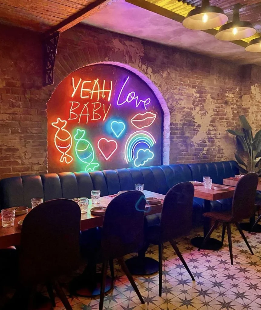 A cozy restaurant with a neon sign displaying "Yeah Baby," "Love," candies, hearts, a rainbow, lips, and clouds on a brick wall.