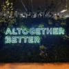 Neon sign displaying "Altogether Better" in bright green letters mounted on a lush greenery wall with a wooden floor below.