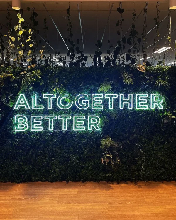 Neon sign displaying "Altogether Better" in bright green letters mounted on a lush greenery wall with a wooden floor below.