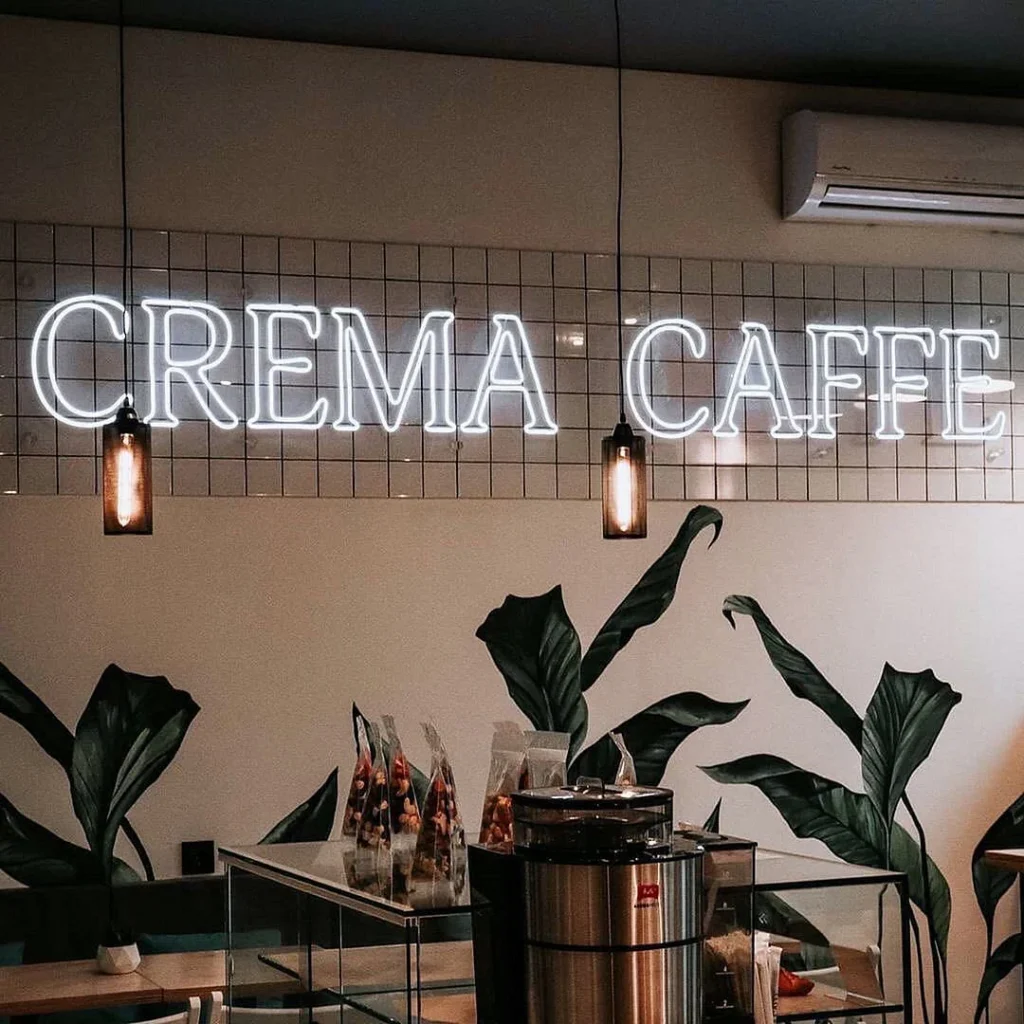 White neon "CREMA CAFFE" sign in a café with Edison bulb lights and green plant decals.