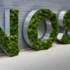 Large metal letters filled with green moss spelling "NOSH" against a gray brick wall.