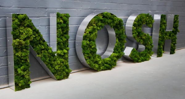 Large metal letters filled with green moss spelling "NOSH" against a gray brick wall.