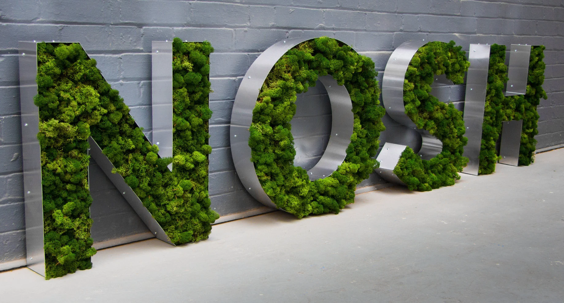 Large metal letters filled with green moss spelling "NOSH" against a gray brick wall.