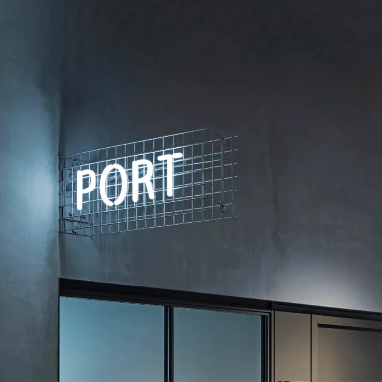 Neon sign with the word "PORT" in white letters encased in a metal grid, mounted on an exterior wall above large glass windows.