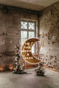 A neon sign in the shape of a crescent moon with the words "TO THE MOON & BACK" illuminated in white light is displayed in front of a rustic, exposed brick wall. The sign is adorned with floral arrangements featuring pink and white flowers at its base and along the side, enhancing its decorative appeal. The backdrop includes a tall window with a view of greenery, adding natural light to the scene. This setup is used for a romantic event or wedding.