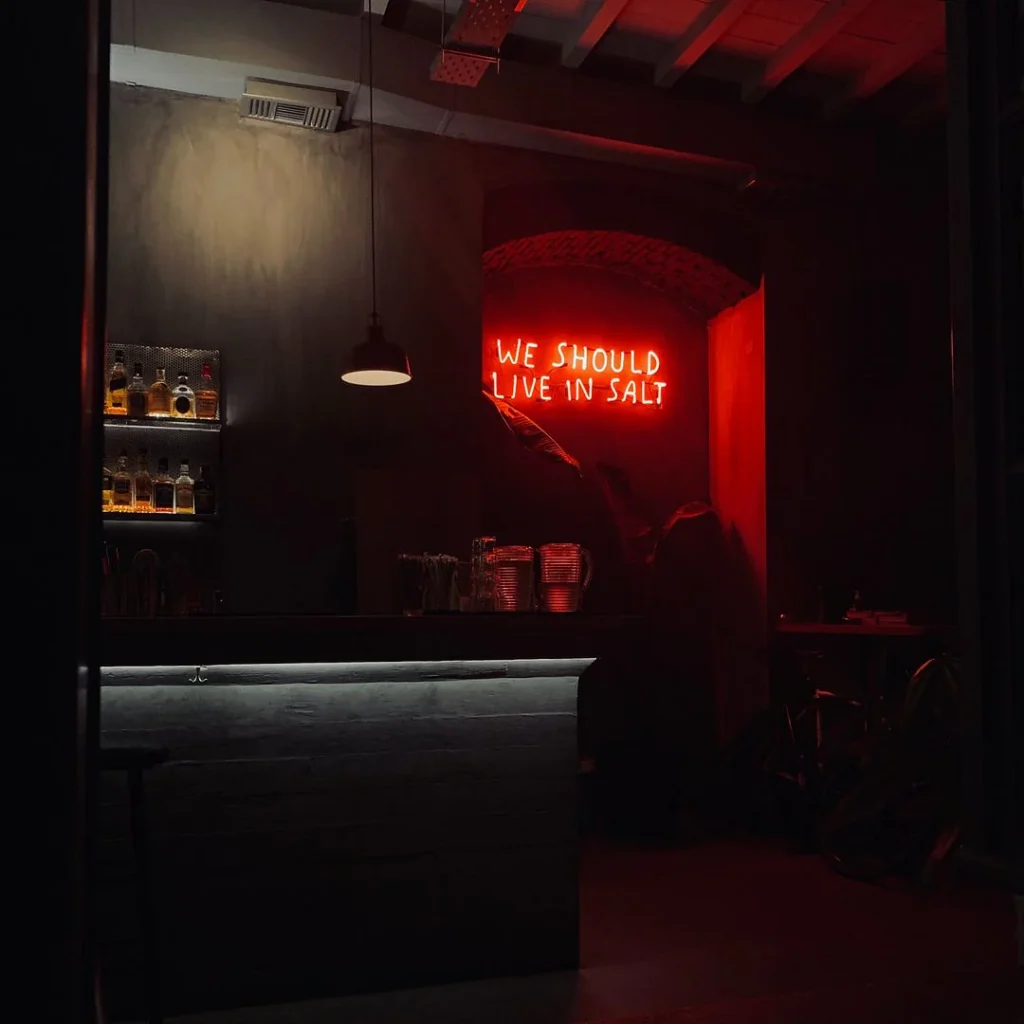 A dimly lit bar featuring a red neon sign that reads "We Should Live In Salt.