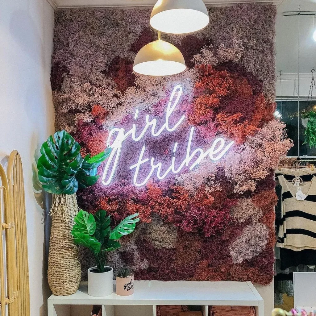 Neon sign saying "girl tribe" in pink cursive letters, mounted on a wall covered with pink and purple flowers, with a plant in a woven basket below.