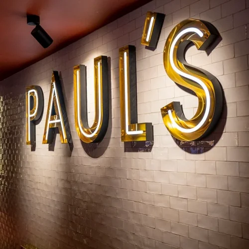 Illuminated neon sign reading "PAUL'S" mounted on a tiled wall inside a restaurant. The sign features large, bold letters with a yellow and white glow, enhancing the upscale ambiance of the venue. The sign is situated above a hallway leading to other areas of the restaurant.