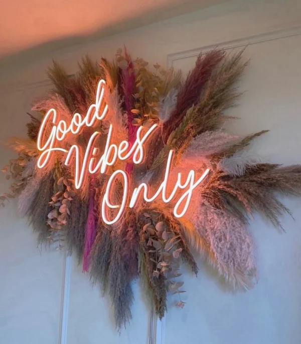 Neon sign reading "Good Vibes Only" in white cursive letters, surrounded by a decorative arrangement of dried flowers in various shades.