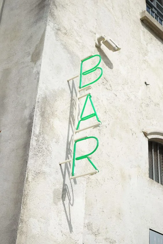 A vertical green neon "BAR" sign mounted on the exterior wall of a building. The sign consists of green neon tubing spelling the word "BAR" in a vertical stack.