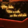 Neon sign in orange cursive letters reading "Hey babe, Take a walk on the wild side" mounted on a lush greenery wall with warm ambient lighting.