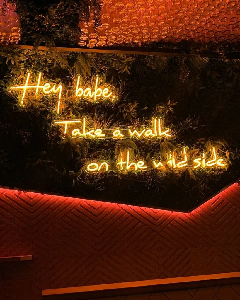 Neon sign in orange cursive letters reading "Hey babe, Take a walk on the wild side" mounted on a lush greenery wall with warm ambient lighting.