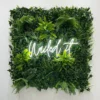 Neon sign saying "Nailed it" in white cursive letters, mounted on a square greenery wall with various green plants.