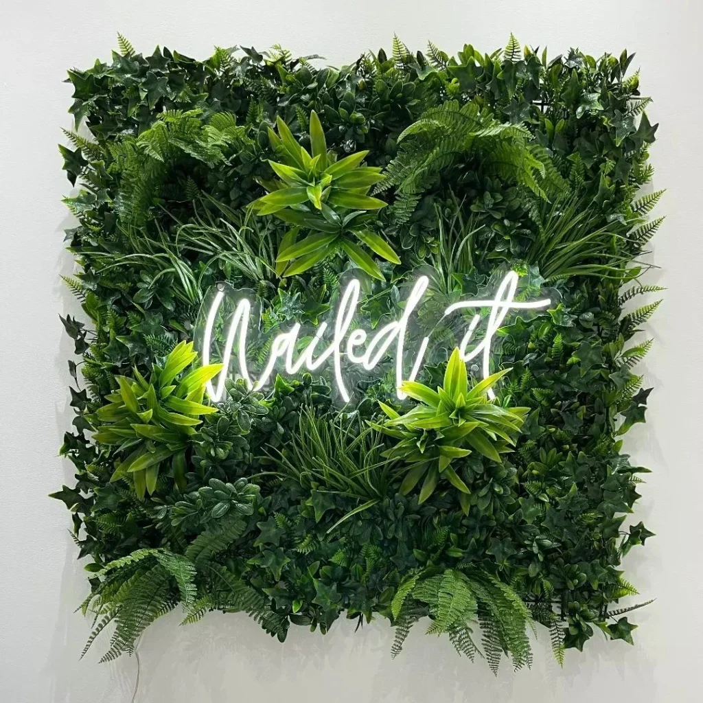 Neon sign saying "Nailed it" in white cursive letters, mounted on a square greenery wall with various green plants.