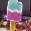 Neon sign of a bitten ice cream popsicle with light blue and pink sections, surrounded by reflective silver and gold balloons.