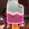 Neon sign of a bitten ice cream popsicle with light blue and pink sections, surrounded by reflective silver and gold balloons.