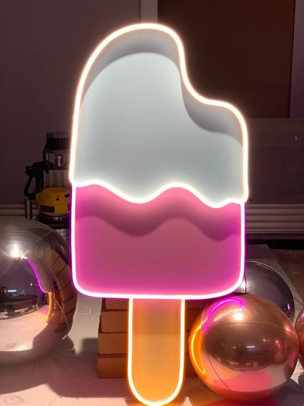 Neon sign of a bitten ice cream popsicle with light blue and pink sections, surrounded by reflective silver and gold balloons.