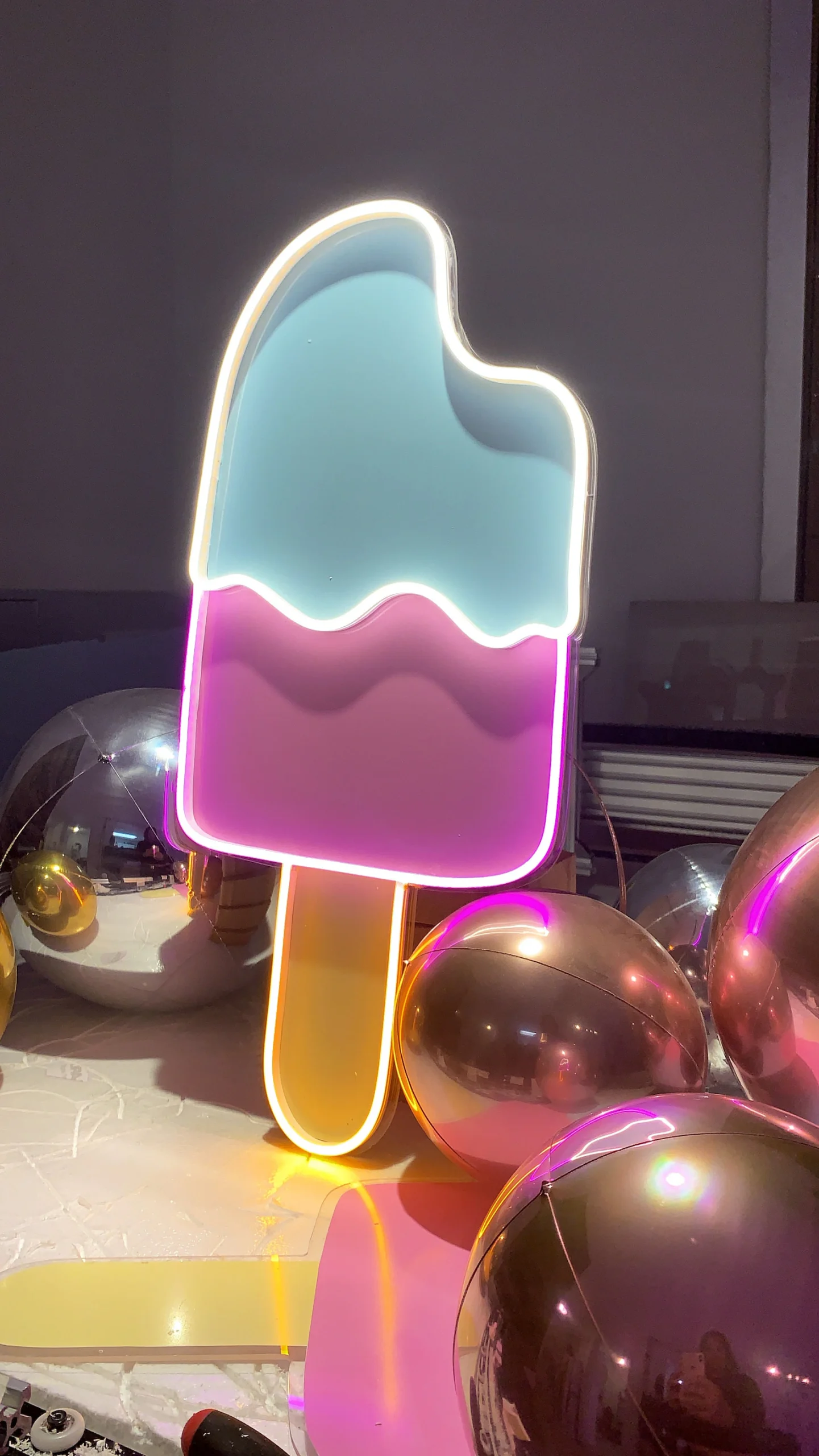 Neon sign of a bitten ice cream popsicle with light blue and pink sections, surrounded by reflective silver and gold balloons.