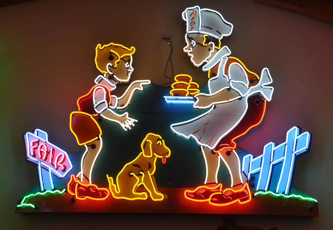 Neon sign featuring a colorful scene of a boy, a chef, and a dog. The boy is pointing towards the chef who is holding a tray of burgers. A sign labeled "Fair" is on the left, and a white fence is on the right.