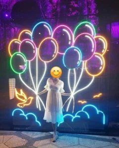 A woman in a white dress stands in front of a neon sign featuring colorful balloons, blue clouds, and a yellow dove against a purple-lit background.