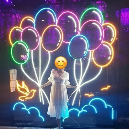 A woman in a white dress stands in front of a neon sign featuring colorful balloons, blue clouds, and a yellow dove against a purple-lit background.