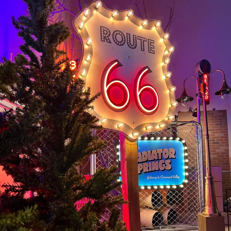 A vibrant display featuring a Route 66 neon sign with a festive Christmas tree on the left and a Radiator Springs sign in the background, creating a nostalgic and retro ambiance.