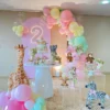 Pastel balloon arch in pink, yellow, and mint green with a neon number "2," plush animal toys, and floral arrangements.