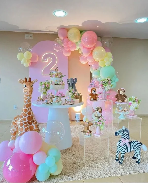 Pastel balloon arch in pink, yellow, and mint green with a neon number "2," plush animal toys, and floral arrangements.